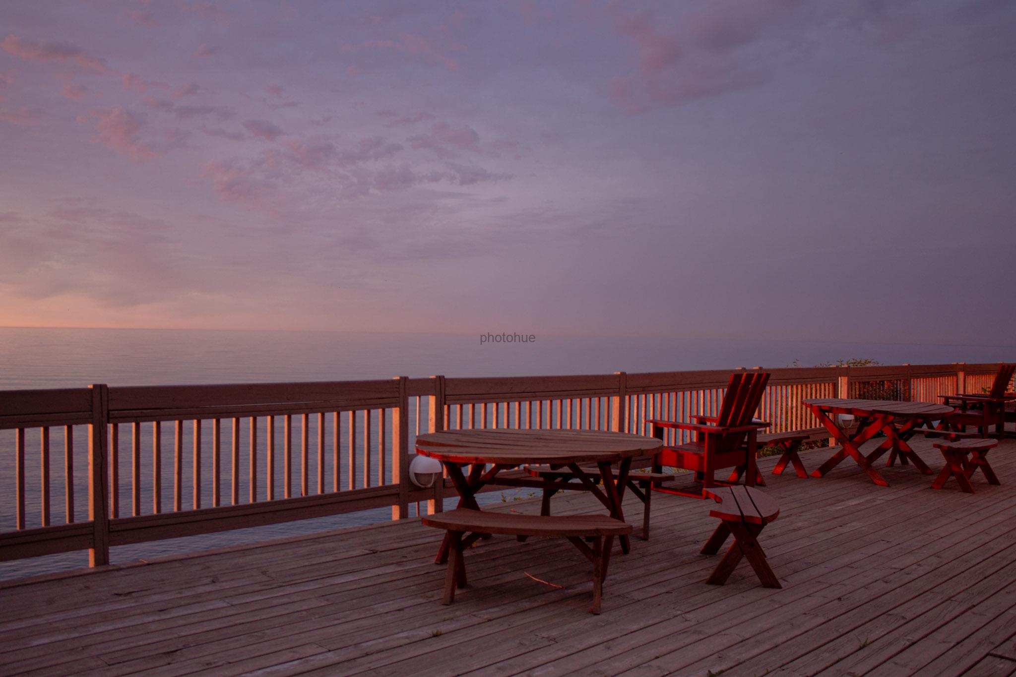 Empty wooden deck with picnic tables and chairs overlooking a calm lake at dawn, under a soft purple and pink sky.